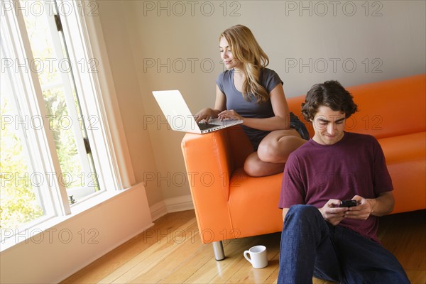 Young couple in living room, using phone and laptop. Photo : Rob Lewine