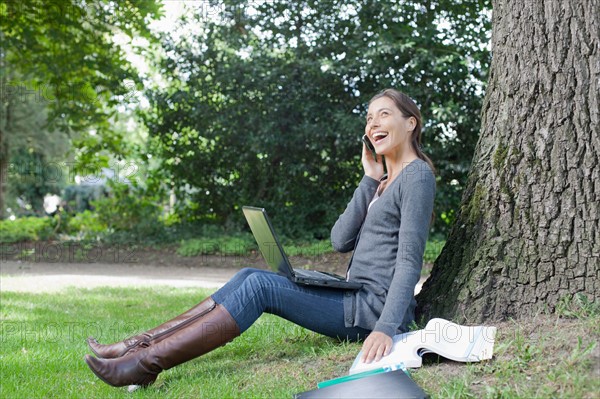 Young woman sitting under tree using laptop and cell phone. Photo : Jan Scherders