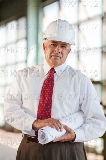 Portrait of senior man wearing tie and hardhat, holding blueprint. Photo: db2stock