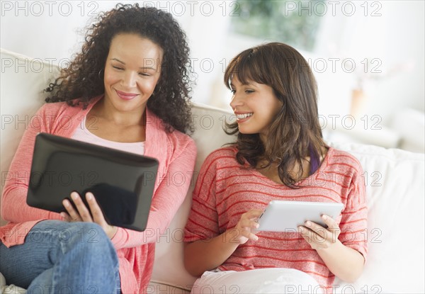 Two women sitting on sofa using digital tablets.