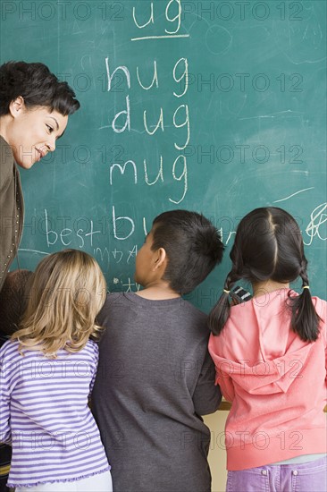 Children (6-7) writing on blackboard with teacher looking at them. Photo : Rob Lewine