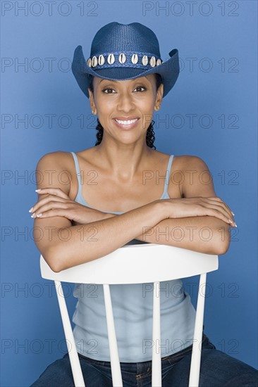 Portrait of mid adult woman sitting on white chair, wearing blue cowboy hat. Photo : Rob Lewine