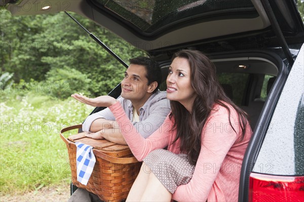 Couple with picnic basket sitting in car trunk. Photo : Tetra Images