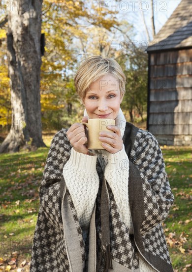 Portrait of smiling woman holding mug in front of cottage house in Autumn. Photo : Tetra Images