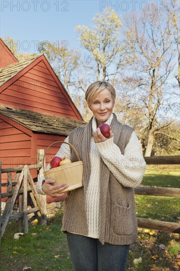 USA, New Jersey, Portrait of smiling woman holding basket with apples in front of cottage house in Autumn. Photo : Tetra Images