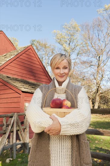 USA, New Jersey, Portrait of smiling woman holding basket with apples in front of cottage house in Autumn. Photo : Tetra Images