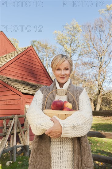 USA, New Jersey, Portrait of smiling woman holding basket with apples in front of cottage house in Autumn. Photo: Tetra Images