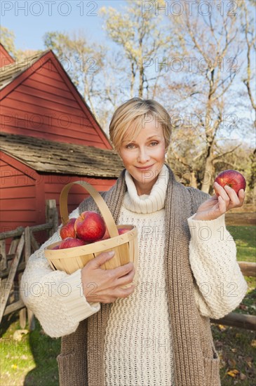 USA, New Jersey, Portrait of smiling woman holding basket with apples in front of cottage house in Autumn. Photo: Tetra Images