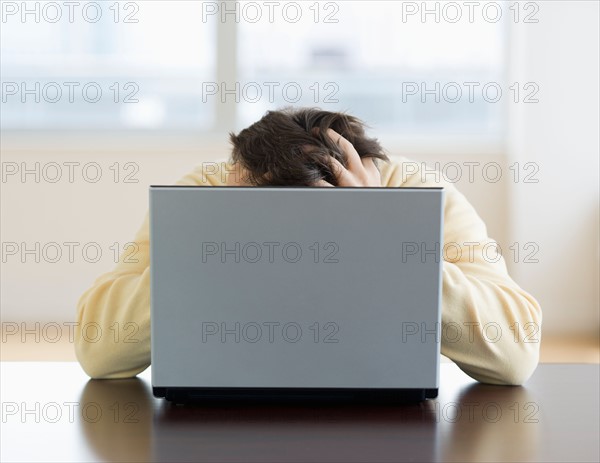 Stressed man sitting at desk behind laptop with head in hands.