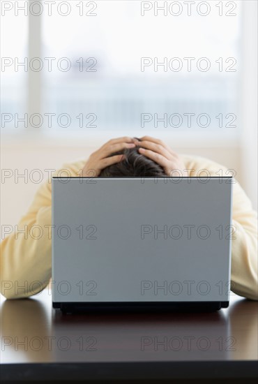 Stressed man sitting at desk behind laptop with head in hands.