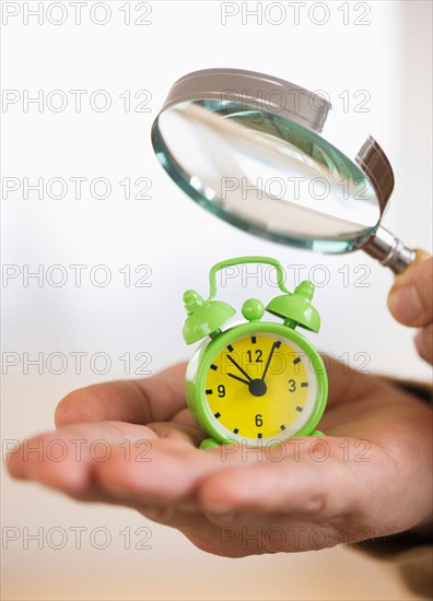 Close up of man's hand holding alarm clock and magnifying glass, studio shot.