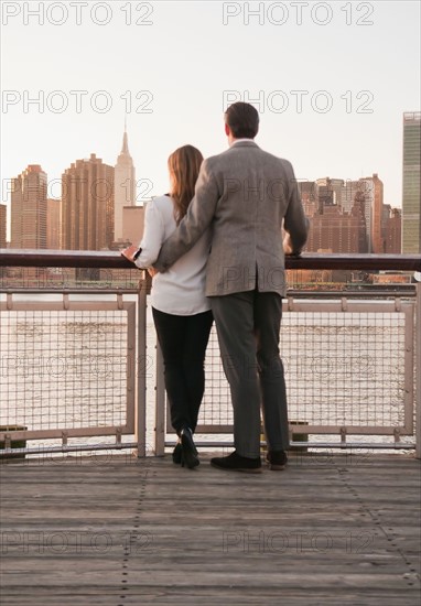USA, New York, Long Island City, Rear view of young couple standing on boardwalk, Manhattan skyline in background. Photo : Daniel Grill