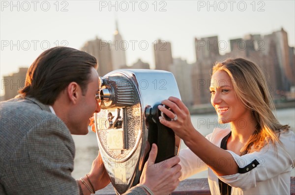 USA, New York, Long Island City, Young couple looking through coin operated binoculars, Manhattan skyline in background. Photo : Daniel Grill