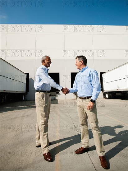 Business men shaking hands outside warehouse.