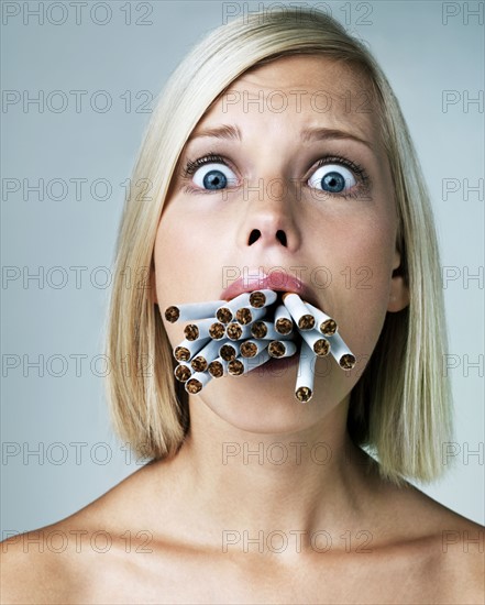 Young woman looking alarmed with mouthful of cigarettes, studio shot. Photo : Yuri Arcurs