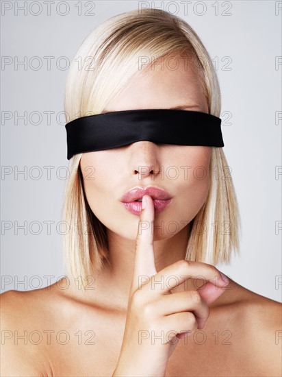 Young woman wearing blindfold with finger on lips, studio shot. Photo : Yuri Arcurs