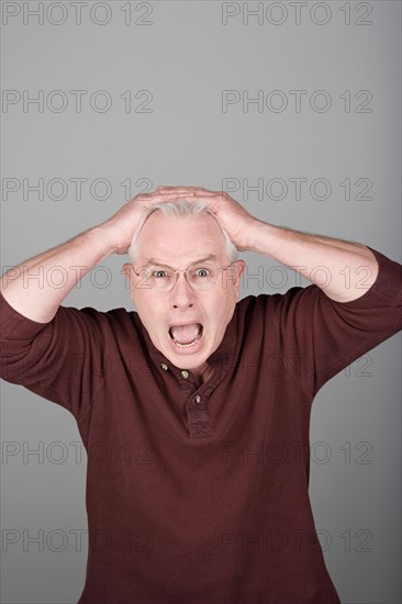 Studio shot portrait of senior man with head in hands and open mouth, waist up. Photo : Rob Lewine