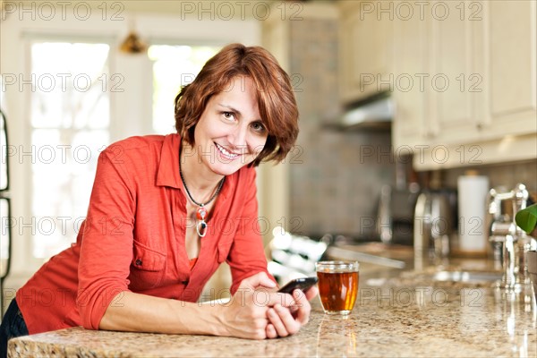 Canada. Ontario, Toronto, Woman in kitchen with glass of tea and cell phone. Photo : Elena Elisseeva