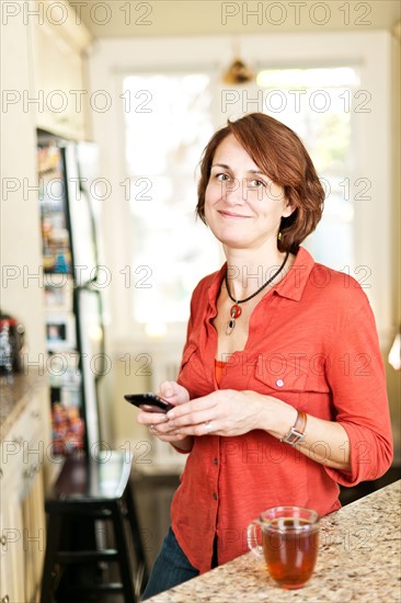 Woman in kitchen with glass of tea and cell phone. Photo : Elena Elisseeva