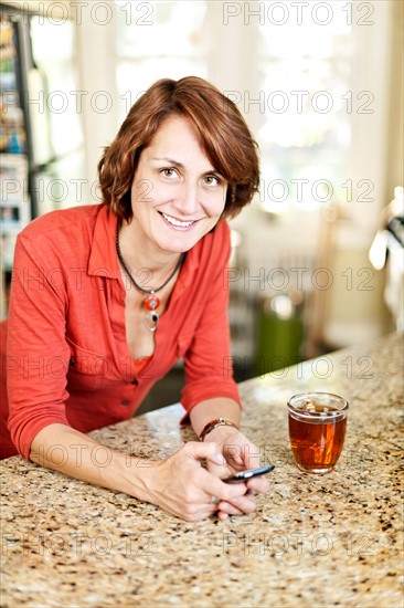 Woman in kitchen with glass of tea and cell phone. Photo : Elena Elisseeva