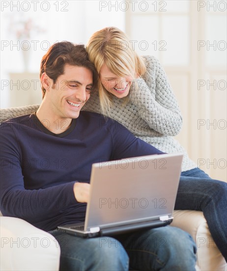Happy couple sitting together on armchair and using laptop . Photo : Daniel Grill