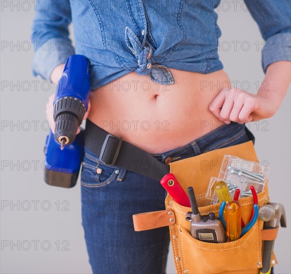 Close up of  young woman wearing tool belt and holding drill, studio shot. Photo : Daniel Grill