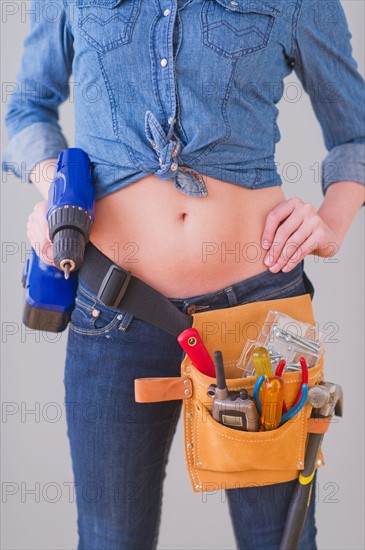 Close up of  young woman wearing tool belt and holding drill, studio shot. Photo : Daniel Grill