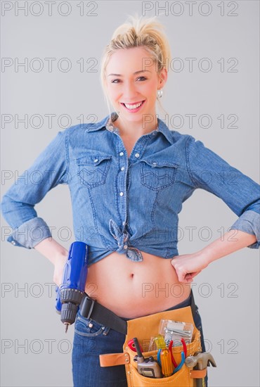 Portrait of young woman wearing tool belt and holding drill, studio shot. Photo : Daniel Grill