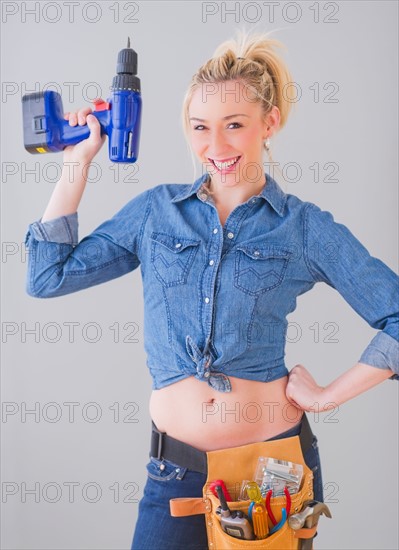 Portrait of young woman wearing tool belt and holding drill, studio shot. Photo : Daniel Grill