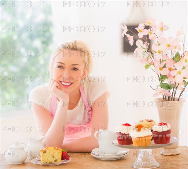 Smiling young woman in apron leaning on table with cakes. Photo : Daniel Grill