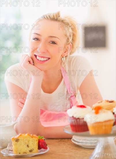 Smiling young woman in apron leaning on table with cakes. Photo : Daniel Grill