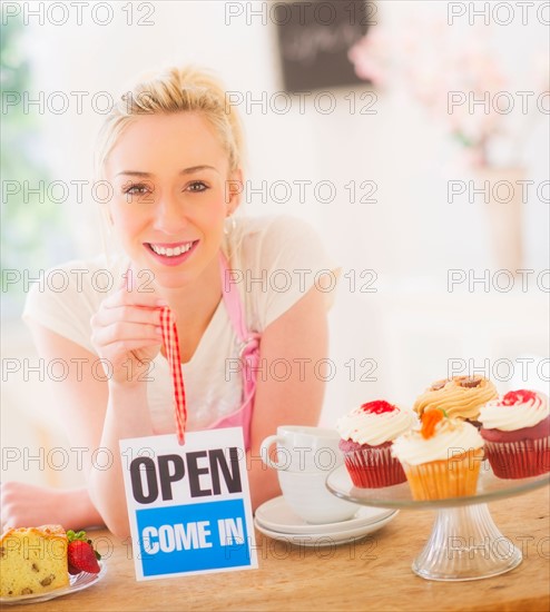 Smiling young woman in apron holding open sign. Photo : Daniel Grill