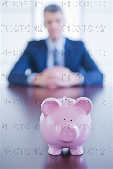 Piggybank on desk, businessman sitting on background. Photo : Daniel Grill