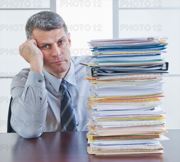 Businessman sitting next to large stack of documents. Photo : Daniel Grill