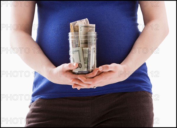 Studio Shot of mid section of pregnant woman holing jar with money. Photo : Mike Kemp