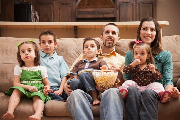 Front view of family with children (18-23 months, 4-5, 6-7, 8-9) sitting on couch watching tv and eating popcorn. Photo : Mike Kemp