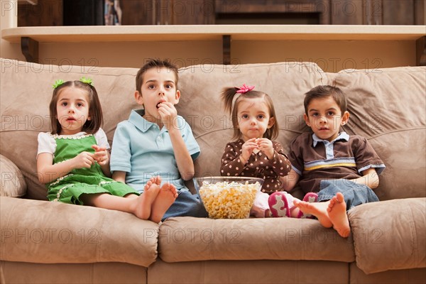 Front view of children (18-23 months, 4-5, 6-7, 8-9) sitting on couch watching tv and eating popcorn. Photo : Mike Kemp