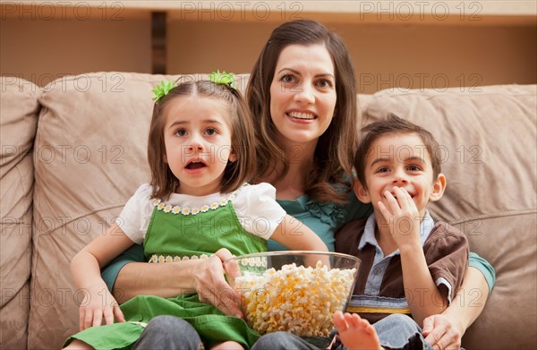 Front view of mother with children (4-5, 6-7) sitting on couch watching tv and eating popcorn. Photo : Mike Kemp