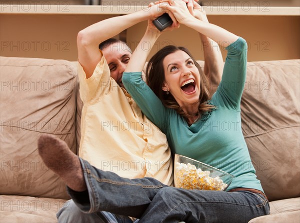 Front view of mid adult couple sitting on couch and fighting for remote control. Photo : Mike Kemp