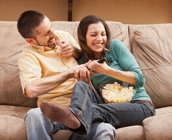 Front view of mid adult couple sitting on couch and fighting for remote control. Photo : Mike Kemp