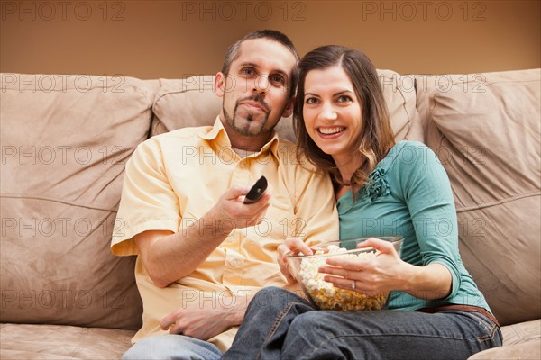 Front view of mid adult couple sitting on couch with remote control and popcorn. Photo : Mike Kemp