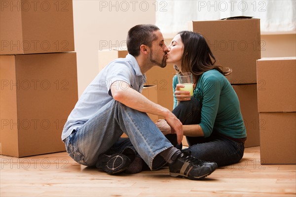 Mid adult couple sitting on floor surrounded by cardboard boxes and kissing. Photo : Mike Kemp