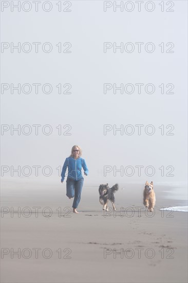 USA, California, Stinson beach. Woman running on sandy beach in fog with her dogs beside her. Photo : Noah Clayton