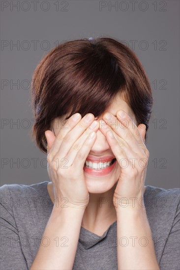 Studio shot of young smiling woman covering face with hands. Photo : Rob Lewine