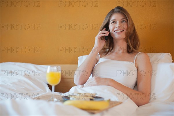 Young woman calling friend while enjoying breakfast in bed. Photo : Mark de Leeuw