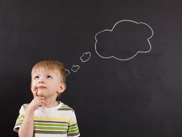 Cute toddler boy (2-3) standing against blackboard with thought bubble written in chalk. Photo : Jessica Peterson