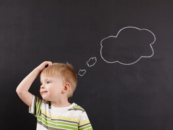 Cute toddler boy (2-3) scratching his head against blackboard with thought bubble written in chalk. Photo : Jessica Peterson