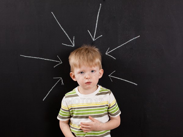 Cute toddler boy (2-3) standing against blackboard with arrows written in chalk pointing at him. Photo : Jessica Peterson