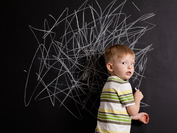 Cute toddler boy (2-3) standing against blackboard with daub written in chalk. Photo : Jessica Peterson