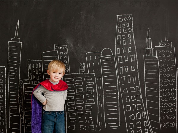 Cute toddler boy (2-3) standing against blackboard with city skyline drawn on it. Photo : Jessica Peterson